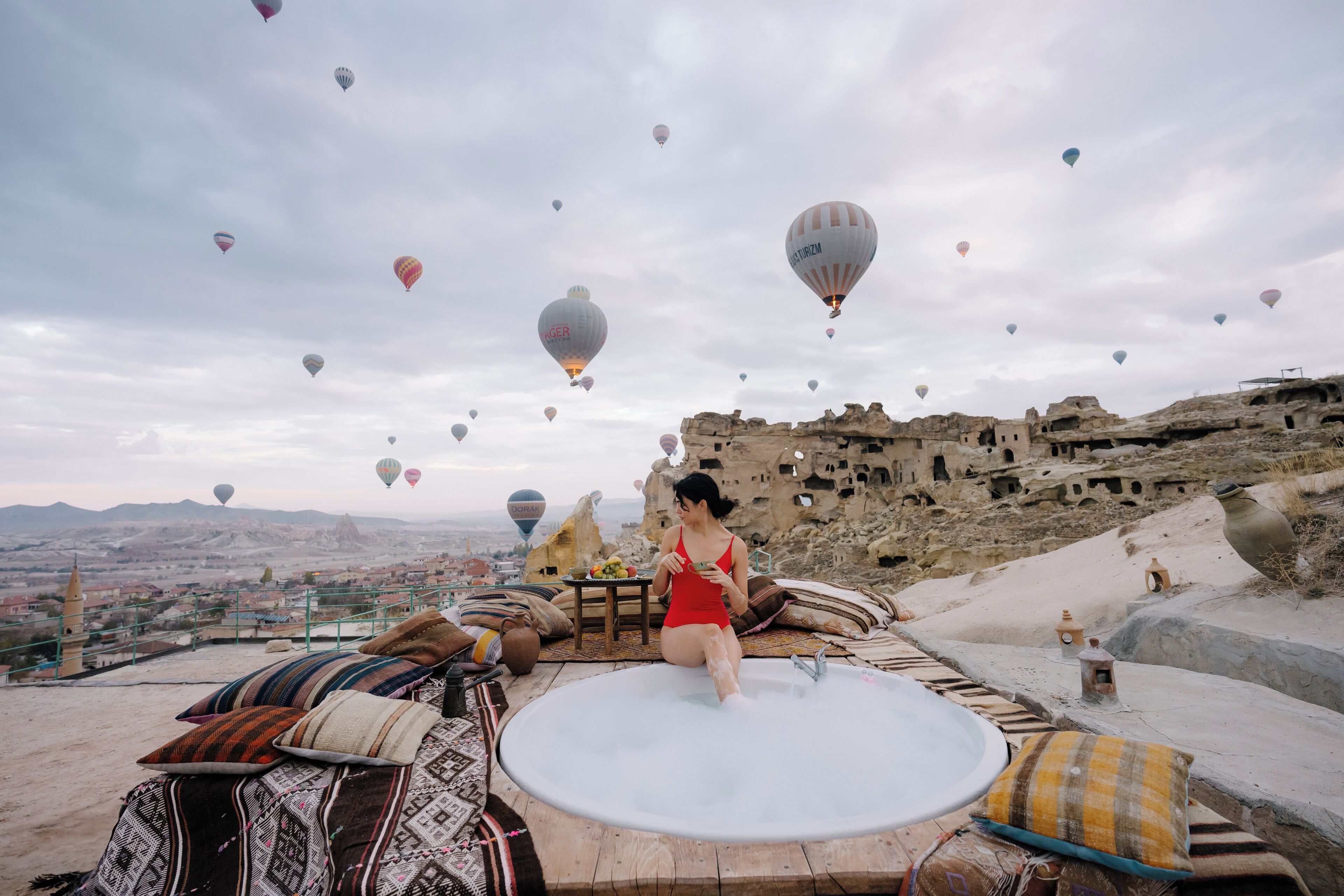 A woman sitting on a hot tub with a view of a city and hot air balloons in the sky.
