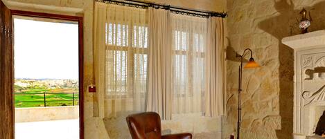A room with a stone wall, a wooden ceiling, a chair, a table with a cup, a floor lamp, and a view of a green landscape through a window.