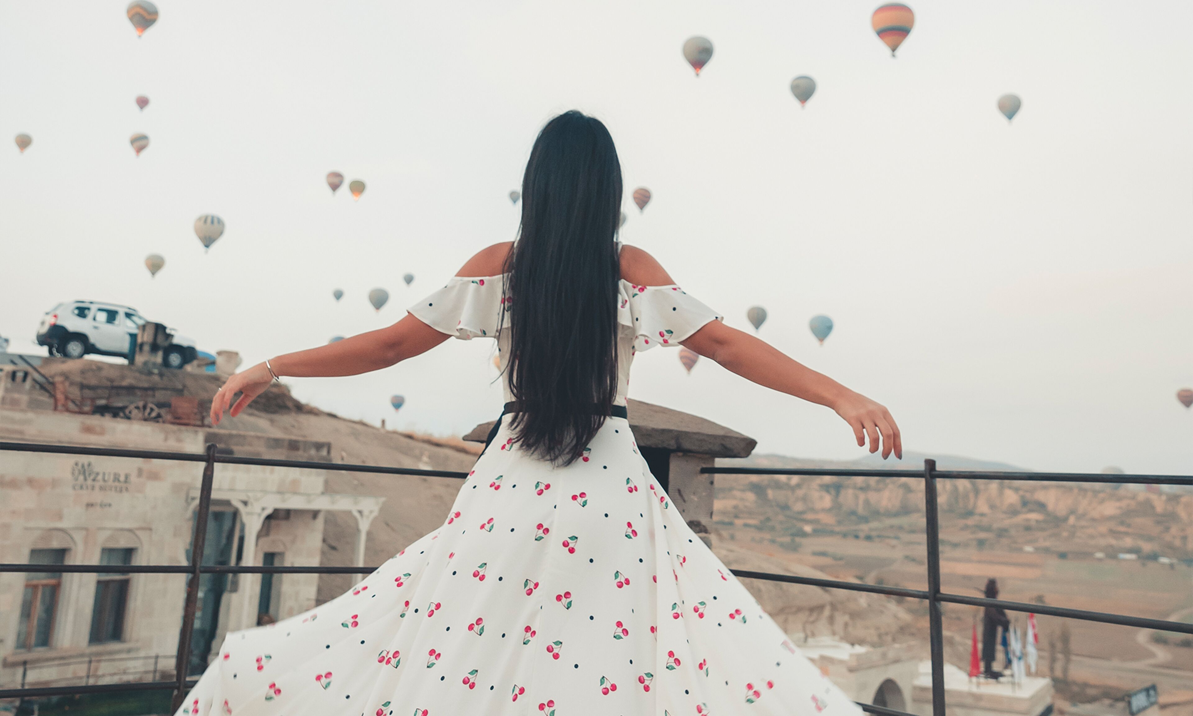 A woman in a white dress with cherry patterns, standing on a balcony with outstretched arms, overlooking a valley with hot air balloons and a vehicle in the background.