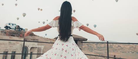 A woman in a white dress with cherry patterns, standing on a balcony with outstretched arms, overlooking a valley with hot air balloons and a vehicle in the background.