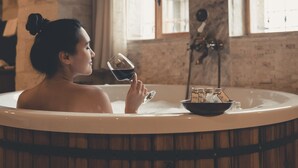 A person in a bathtub with a ship model and bottles, in a room with exposed brick walls and large windows. - Seki Cave Hotel, Wake Up to Hot Air Balloons (Avanos)