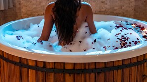 A person sitting in a wooden bathtub filled with bubbles and rose petals, with a view of the sea through large windows. - Seki Cave Hotel, Wake Up to Hot Air Balloons (Avanos)