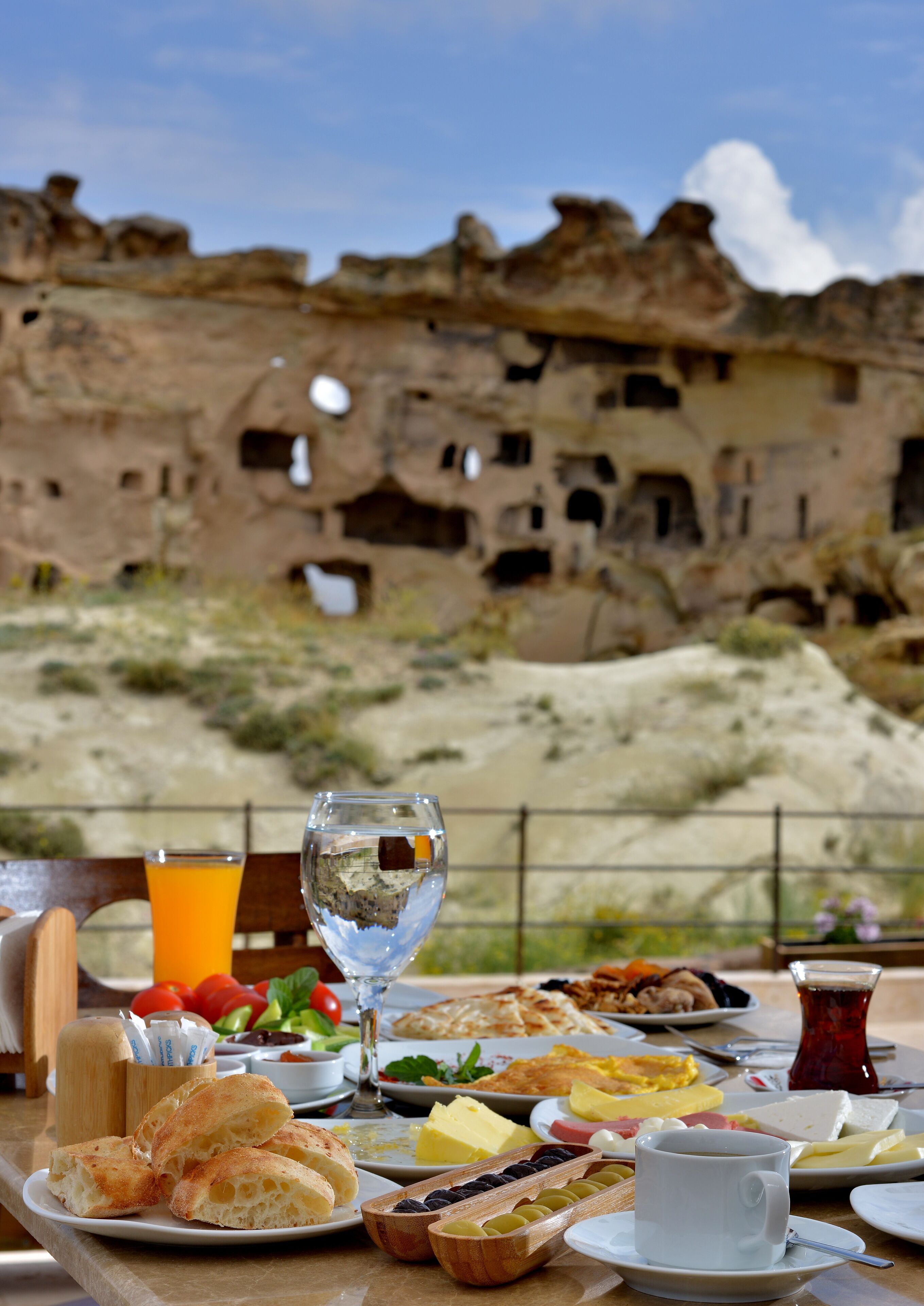 A table set with breakfast items, including bread, cheese, and fruit, with a view of ancient rock formations in the background.
