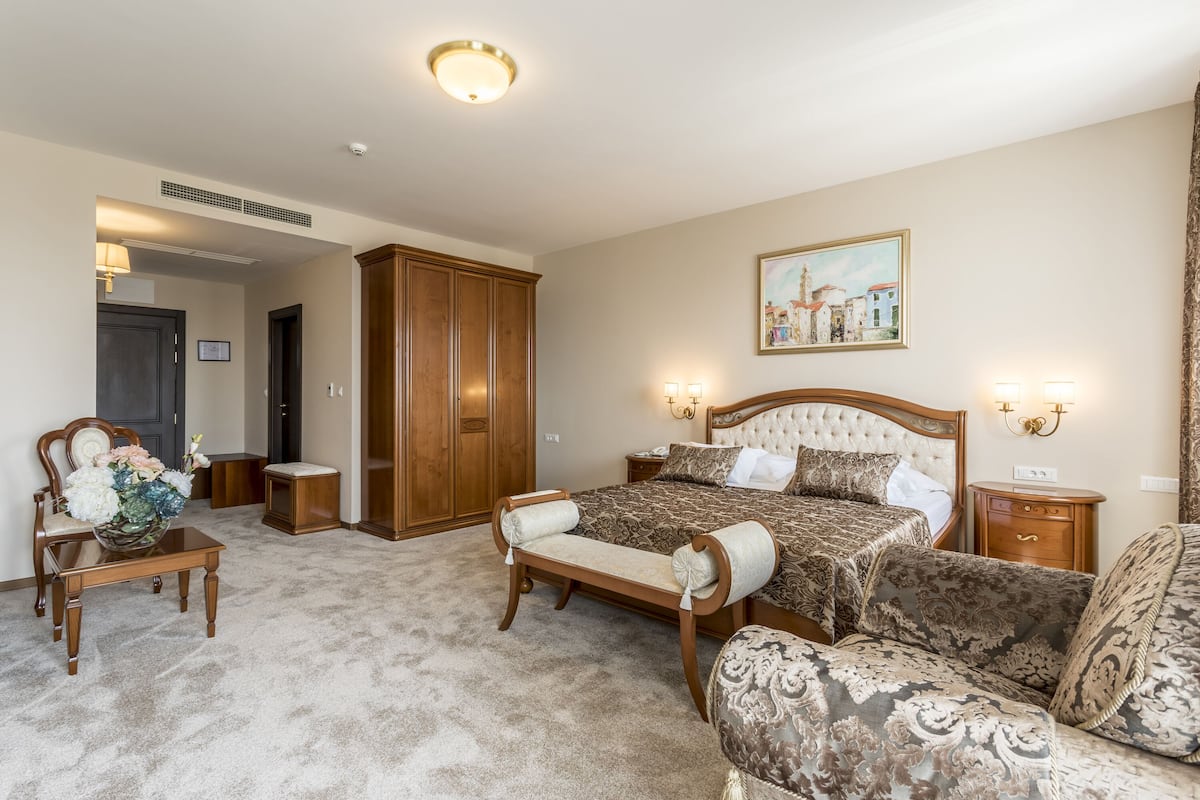 A bedroom with a large bed, a wooden wardrobe, a bench, and a small table with a vase of flowers.