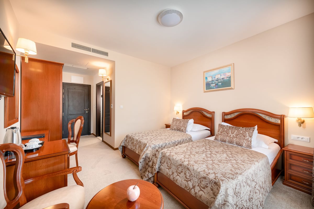 A hotel room with two beds, a desk with a chair, a small table, and a wall-mounted lamp.