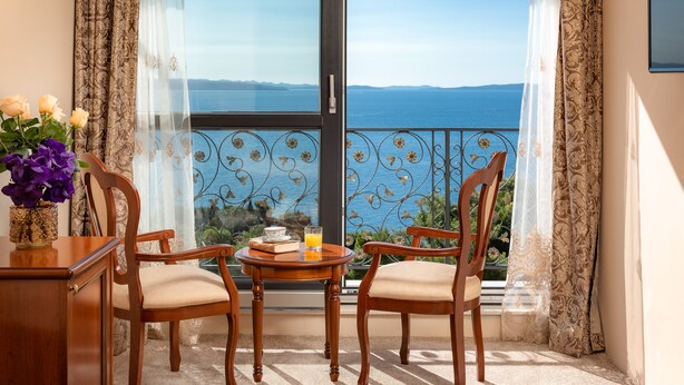 Un balcon avec deux chaises en bois, une petite table ronde sur laquelle se trouve un verre de jus d’orange et un vase rempli de fleurs.
