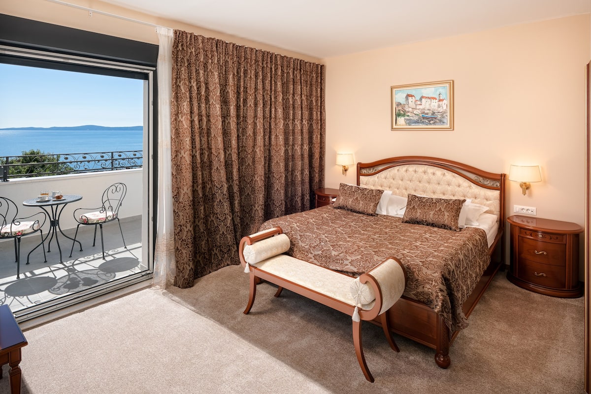 A hotel room with a bed, a bench, a nightstand, a table with chairs, and a view of the sea through a large window.