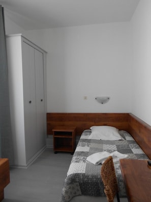 A small, neatly arranged bedroom with a single bed, a wooden nightstand, a wardrobe, and a wall-mounted light fixture. - Hôtel Du Jura (Audincourt)