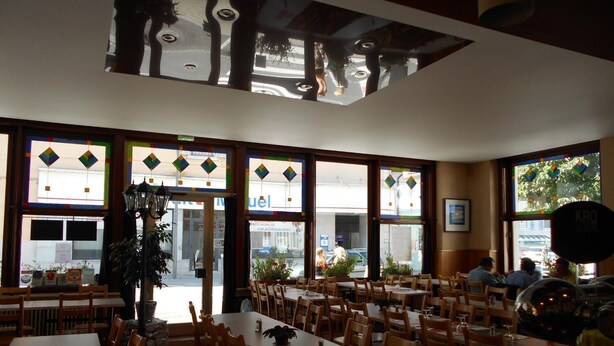 Un restaurant doté de nombreuses tables et chaises, de grandes fenêtres ornées de vitraux et d’un plafond au motif géométrique.