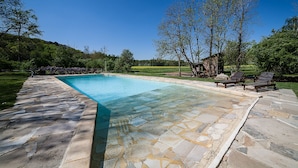 Une piscine rectangulaire revêtue de carreaux de pierre, entourée d’une pelouse et d’arbres.