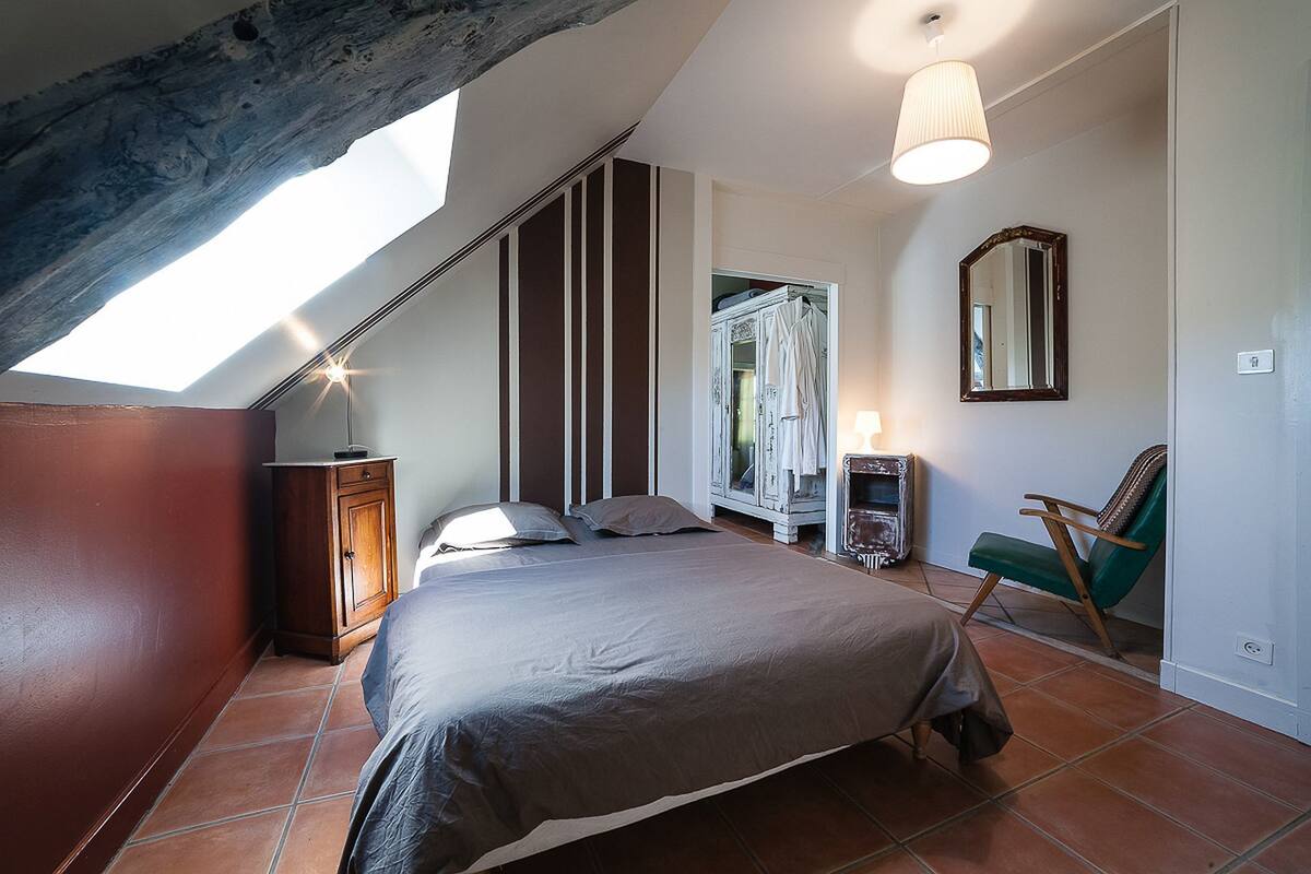 Een slaapkamer met een schuine kap, een bed, een stoel, een kledingkast en een spiegel.