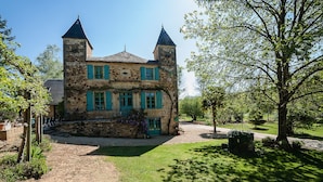 Un bâtiment en pierre doté de deux tours et de volets verts, entouré d’une pelouse bien entretenue et d’arbres.