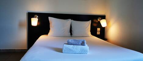 Una cama bien hecha con sábanas blancas, dos almohadas y una toalla doblada. Hay dos lámparas fijadas a la pared a cada lado de la cama.