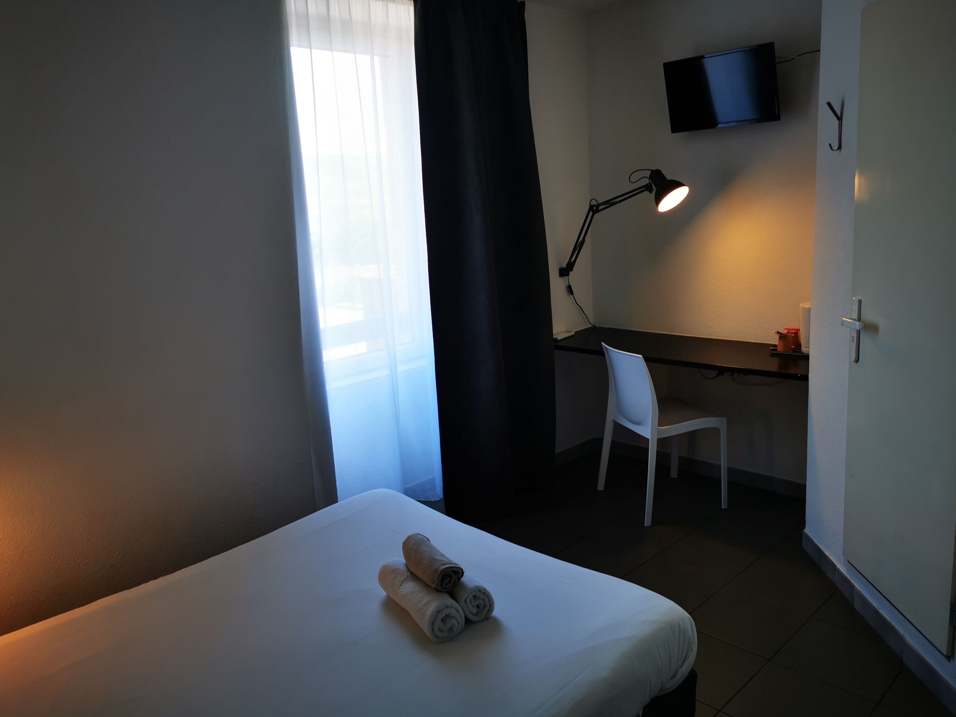 A hotel room with a bed, a desk, a chair, a desk lamp, and a TV mounted on the wall.