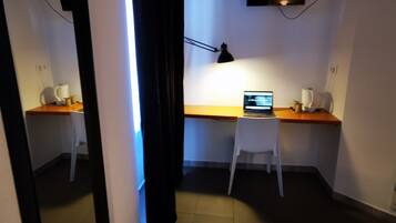 Une petite pièce faiblement éclairée, avec un bureau, une chaise et un ordinateur portable. Un téléviseur à écran plat est fixé au mur, une lampe se trouve sur le bureau, et il y a aussi une machine à café.