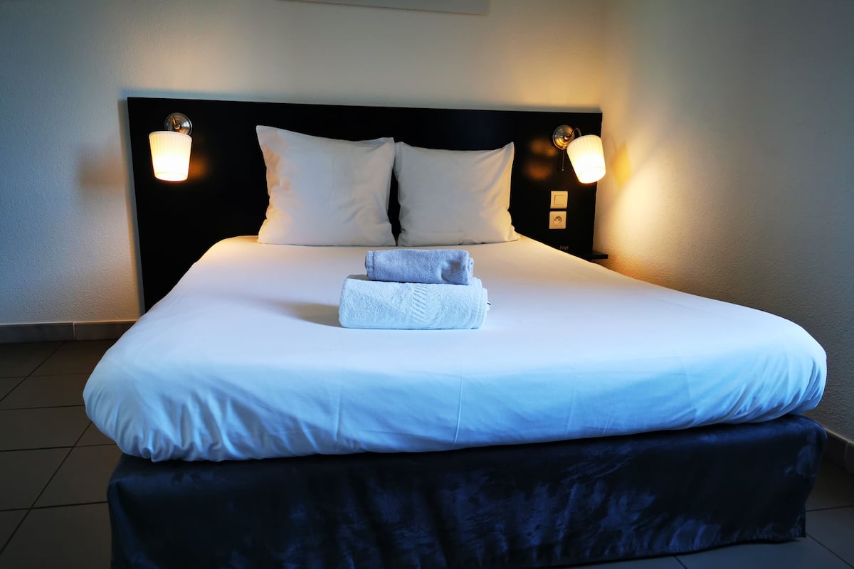 A neatly made bed with white linens and two pillows, a bedside table with a lamp, and a dark-colored ottoman.