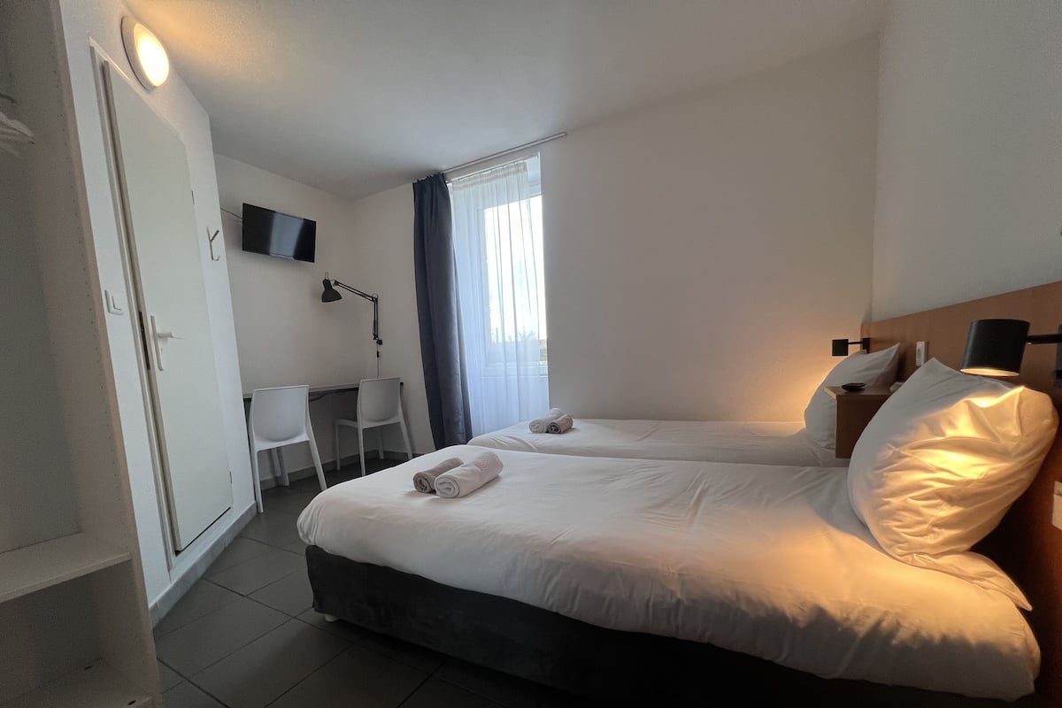 A hotel room with two beds, a desk, a chair, a TV, and a window with curtains.