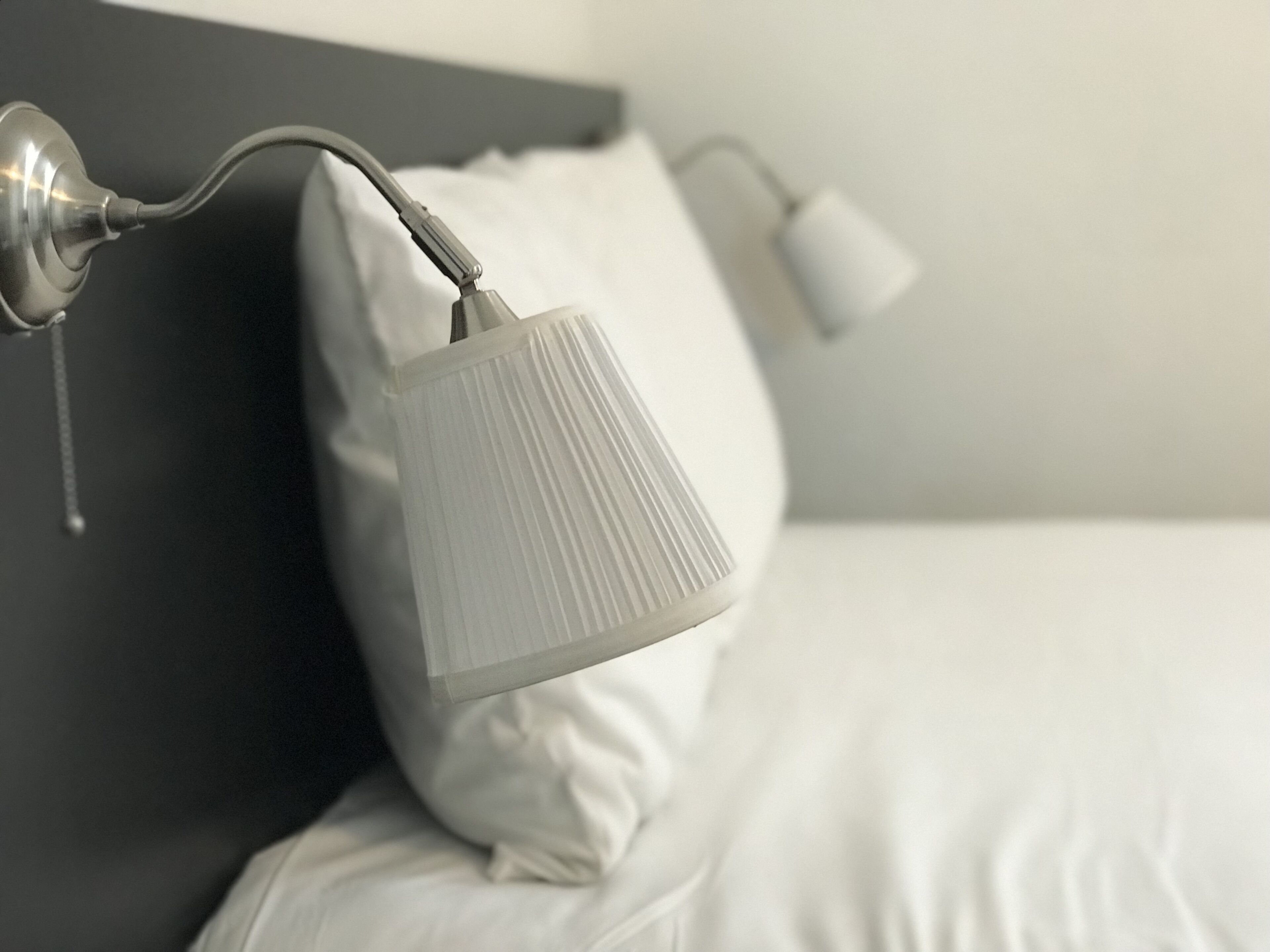 A bed with white bedding, a gray headboard, and a wall-mounted lamp.