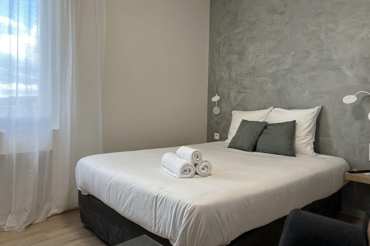 A modern hotel room with a large bed, grey textured wall, and a window with sheer curtains.