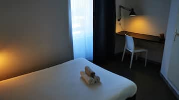A hotel room with a bed, a desk with a chair, a desk lamp, and a TV mounted on the wall.