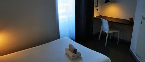 A hotel room with a bed, a desk with a chair, a desk lamp, and a TV mounted on the wall.