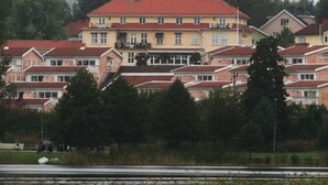 A multi-story residential building with a red-tiled roof, surrounded by trees and a body of water. - Hotell Dalsland (Ed)