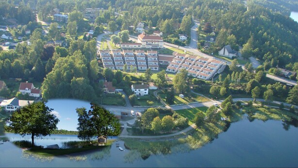 An aerial view of a residential area with a large building complex, a lake, and surrounding greenery. - Hotell Dalsland (Ed)