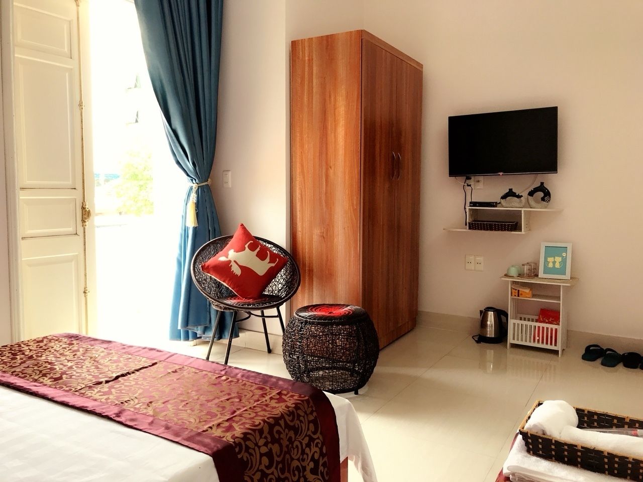A bedroom with a bed, a wooden wardrobe, a television, a chair with a red cushion, a small round stool, and a white shelf with items.