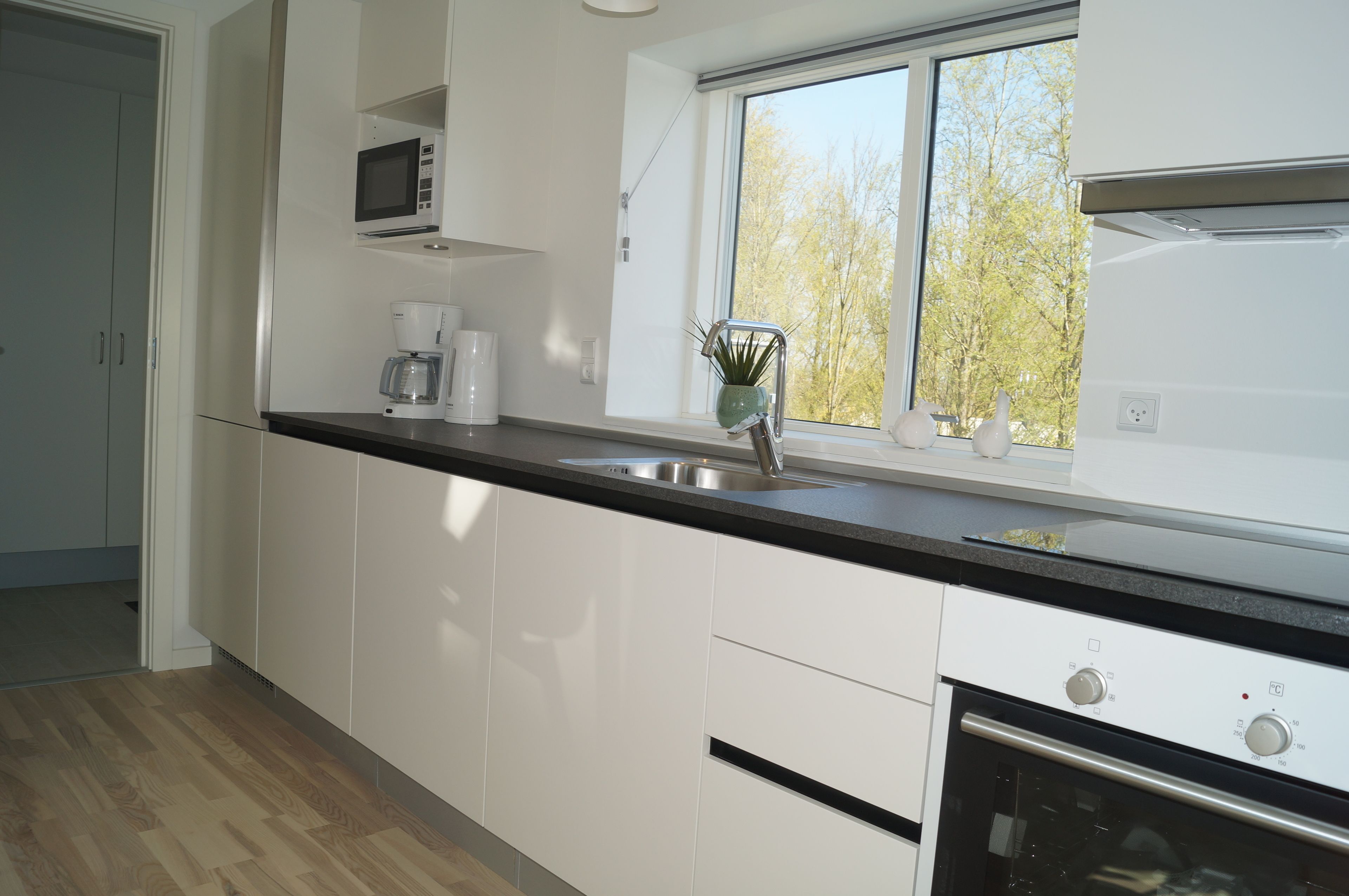 A modern kitchen with white cabinets, a black countertop, a stainless steel sink, a microwave, and a window with a view of trees.