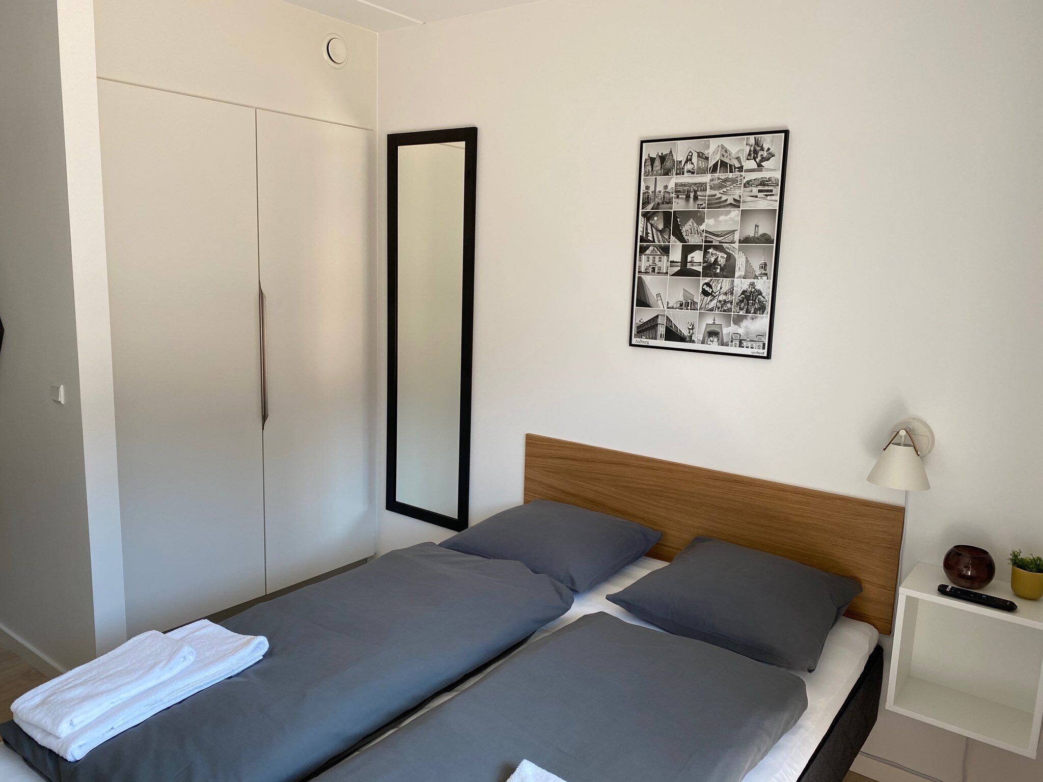 A bedroom with two single beds, a large mirror, a wall-mounted photo collage, a bedside table with a lamp, and a wardrobe.