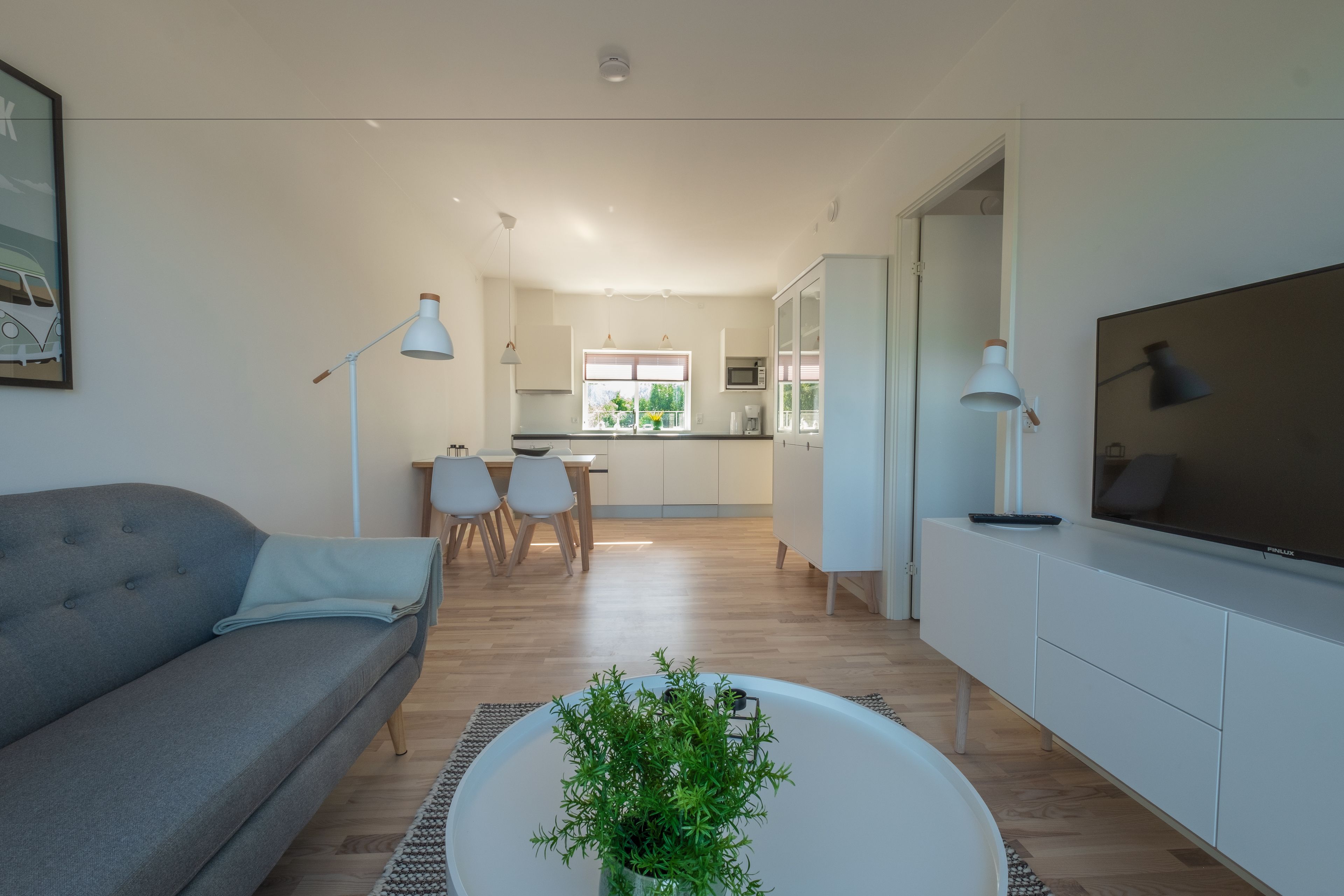 Una sala de estar moderna con un sofá, una mesa de centro y un televisor. Se observa un comedor con sillas y una mesa, y una cocina al fondo.