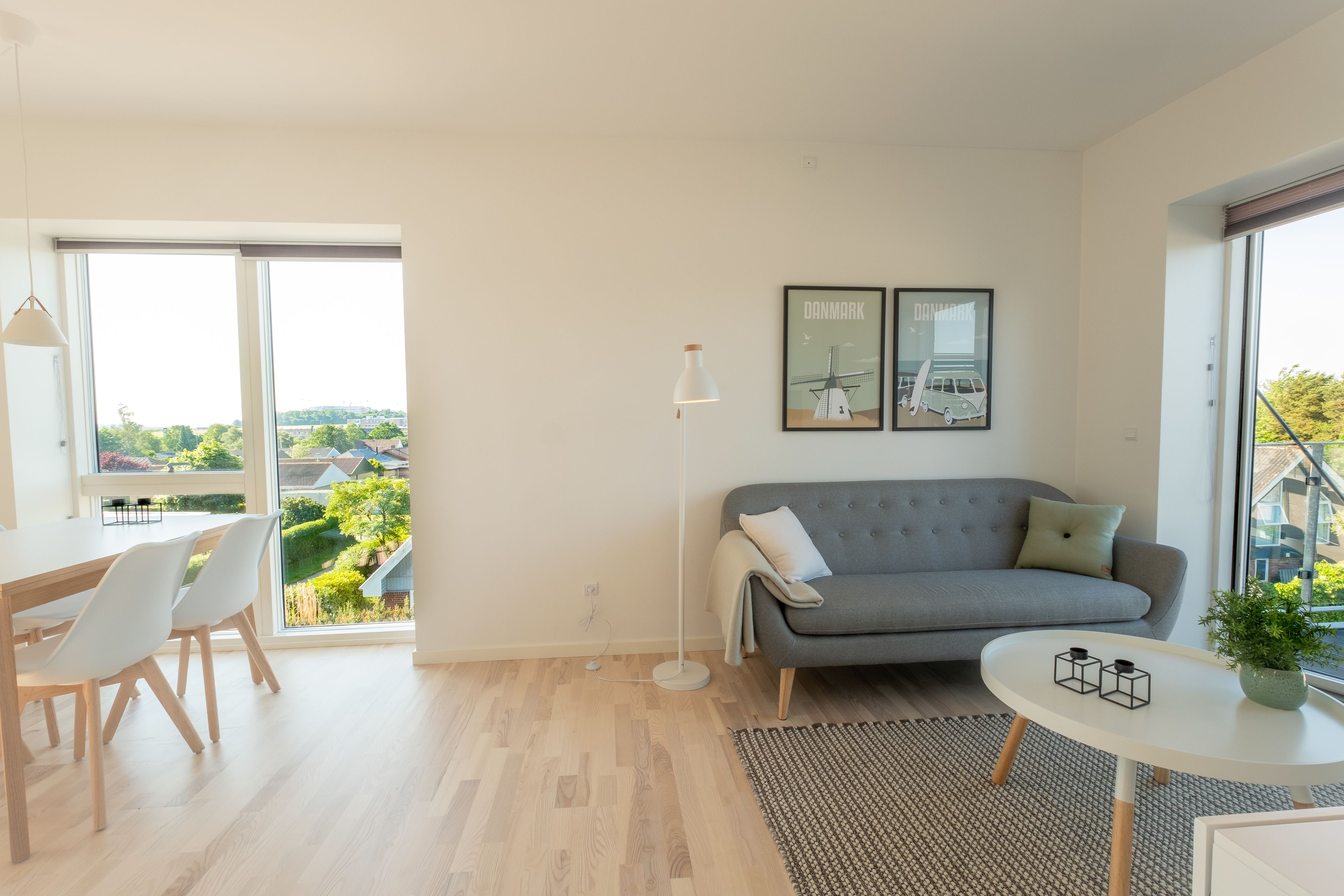 Uma sala de estar moderna com um sofá cinzento, uma mesa redonda branca e uma área de jantar com mesa de madeira e cadeiras brancas. Há duas quadros nas paredes, um abajur e vista para o verde lá fora.