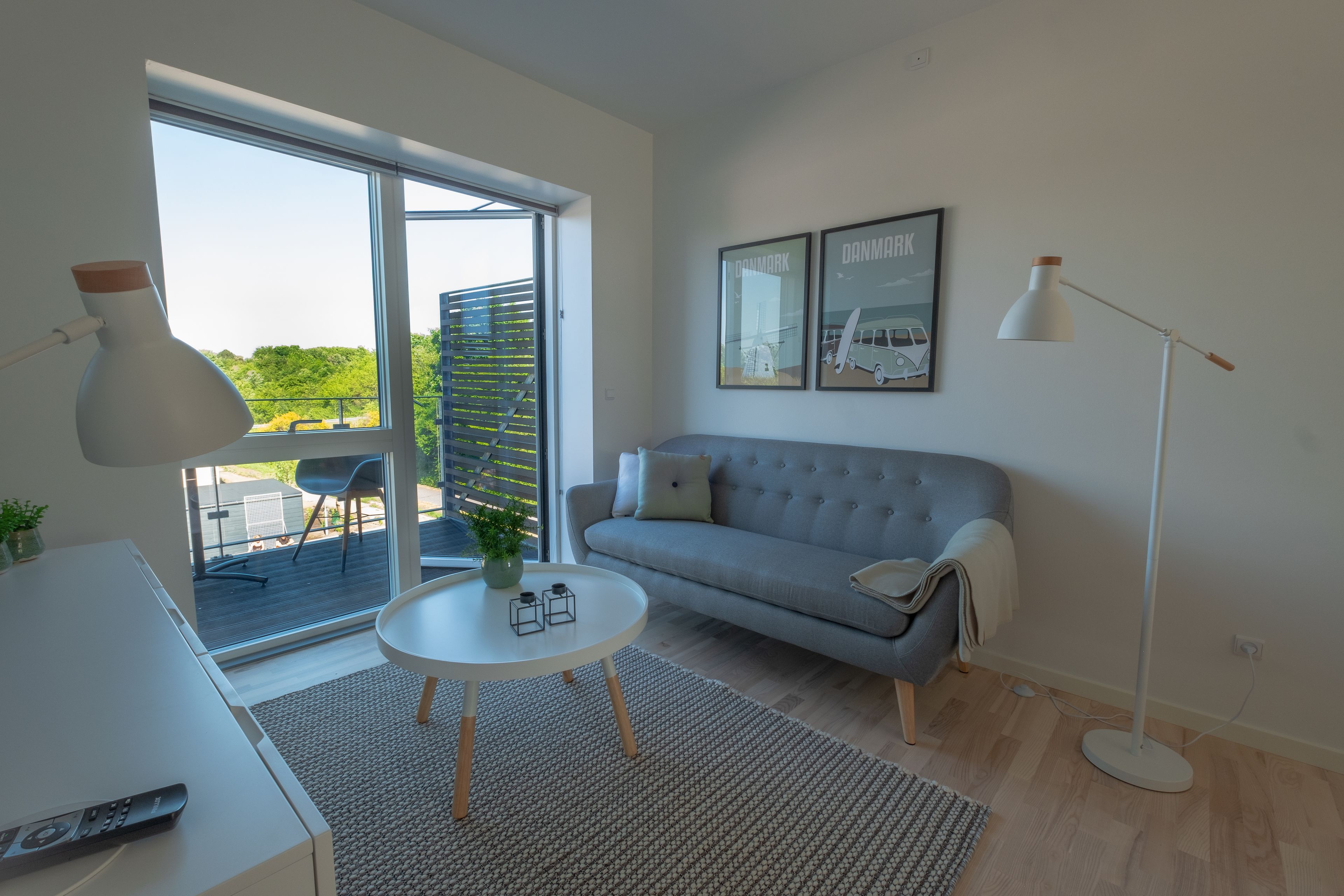 Un salon moderne avec un canapé gris, une table basse ronde, un lampadaire et une grande fenêtre donnant sur un paysage verdoyant et des bâtiments.
