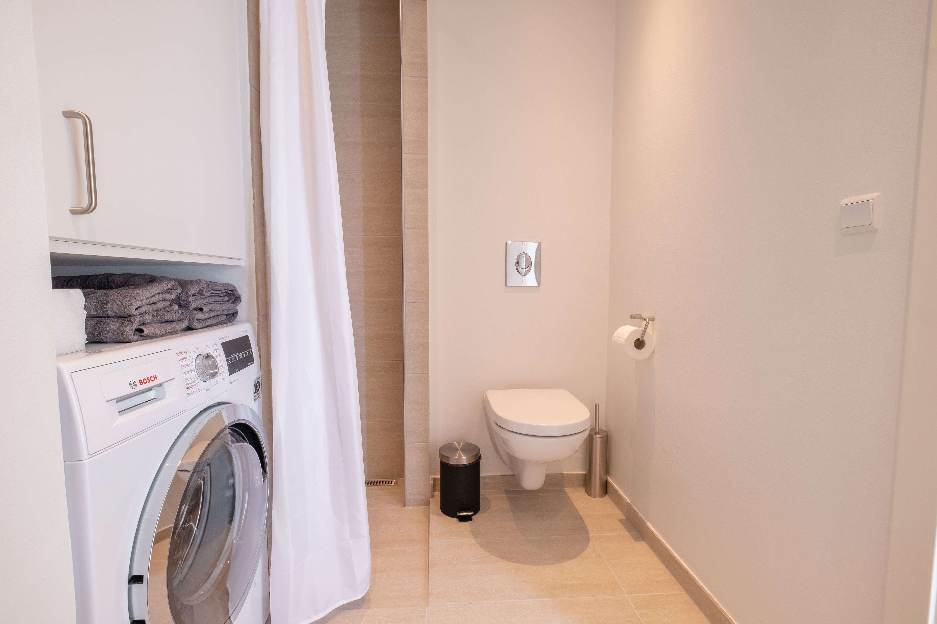 A compact bathroom with a washing machine, toilet, and trash bin.