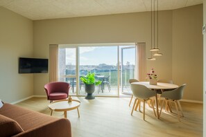 Ein modernes Wohnzimmer mit Fernseher, einer Couch, einem Esstisch mit Stühlen, einer Topfpflanze und einem Balkon mit Aussicht.