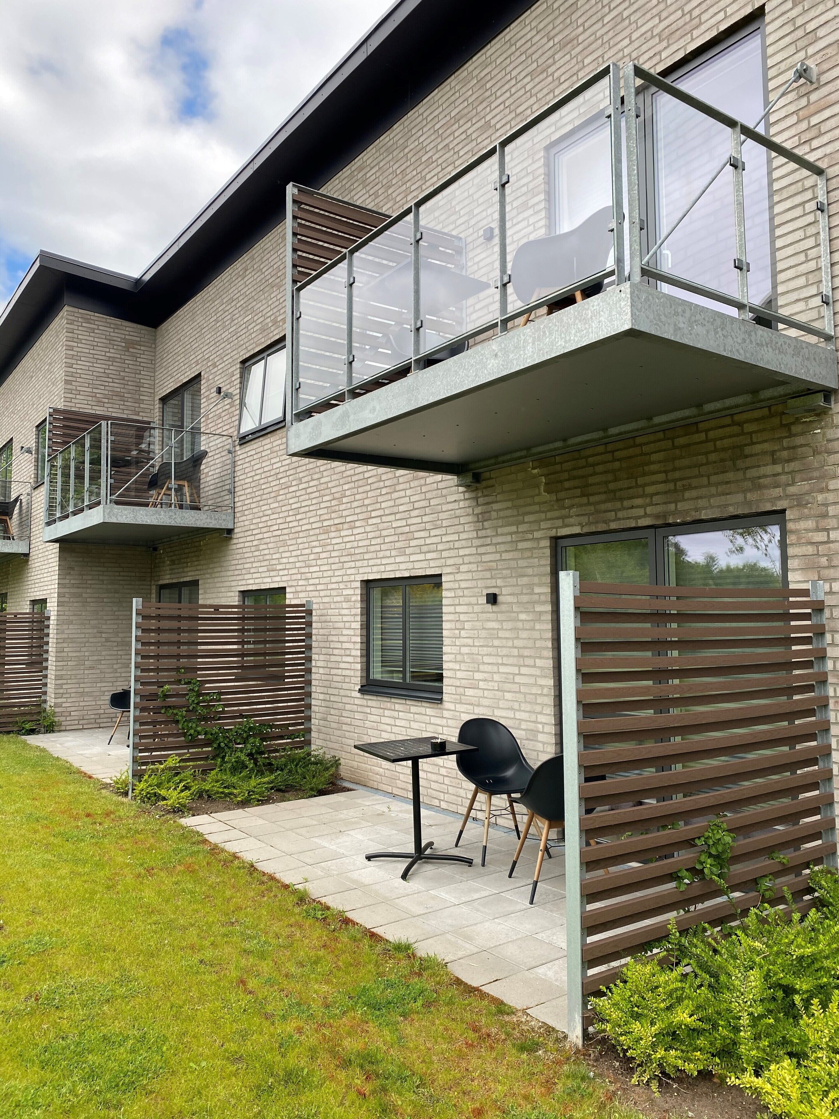 Un immeuble d’appartements moderne doté d’un balcon, d’un petit espace extérieur aménagé avec une table et des chaises, et d’une clôture en bois.