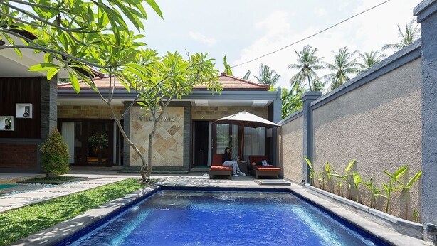 A swimming pool with a tiled deck, a covered seating area with cushions, and a building with a tiled roof and a stone facade. - Villa Twin Lovina (Banjar)