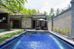 A swimming pool with a tiled deck, a covered seating area with cushions, and a building with a tiled roof and a stone facade. - Villa Twin Lovina (Banjar)