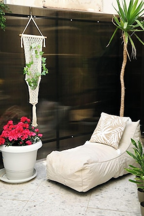 A cozy outdoor seating area with a cushioned chair, a macrame hanging planter with greenery, and a potted plant with red flowers. - My Athens House (Athens)