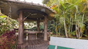 A wooden gazebo with a circular platform and a bench, surrounded by lush tropical plants and a tiled wall. - GuestHouser 4 BHK Bungalow 7283 (Mawal)