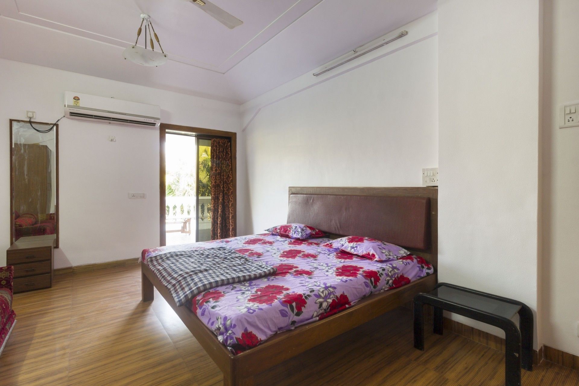 A bedroom with a wooden bed, a small bedside table, a mirror, and a sliding door to a balcony.