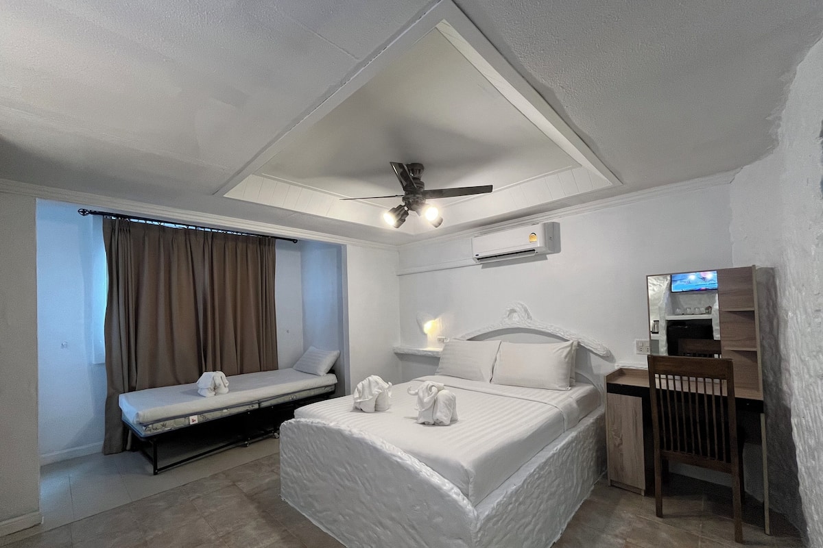 A hotel room with a bed, a desk, a chair, a ceiling fan, an air conditioning unit, and a window with curtains.