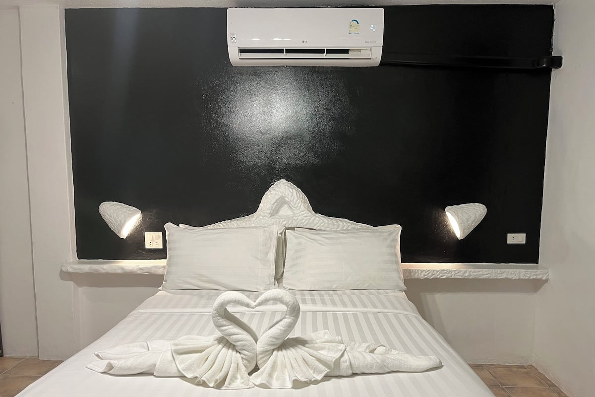 A neatly made bed with a heart-shaped towel arrangement, a black headboard, and an air conditioning unit above.