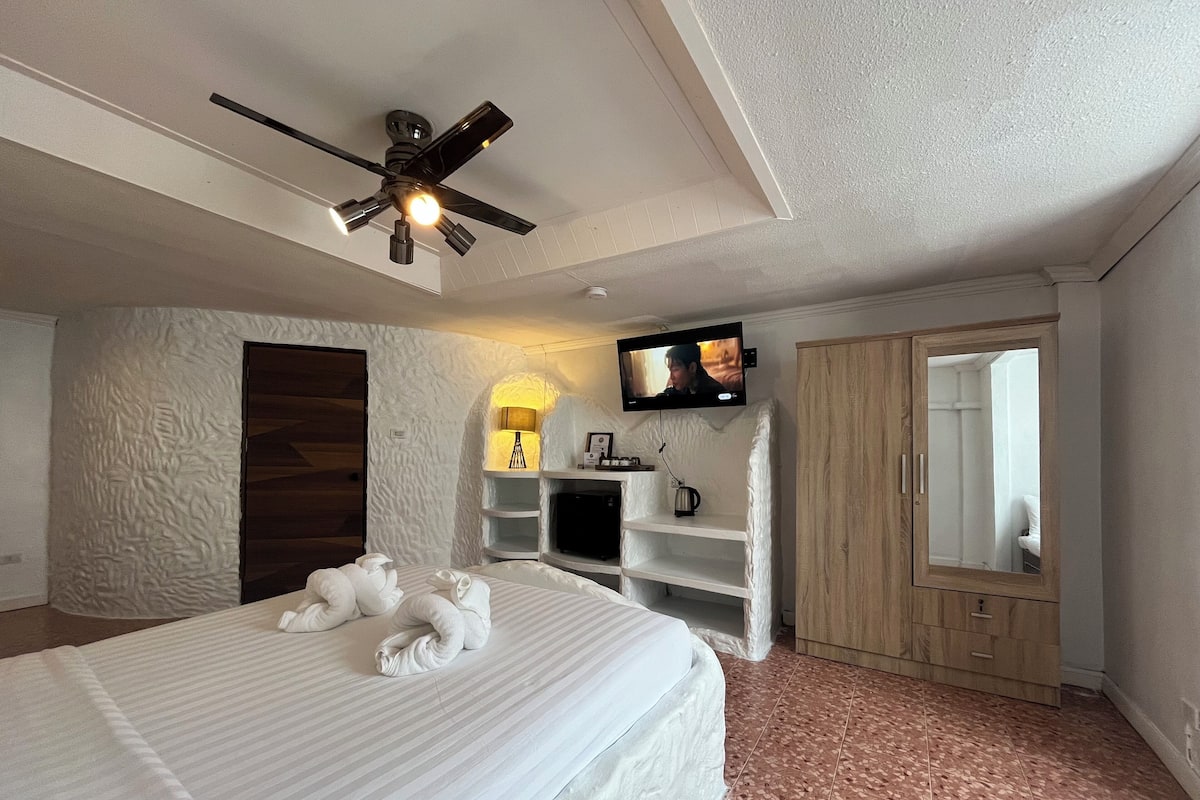 A hotel room with a bed, a ceiling fan, a TV mounted on the wall, a wooden wardrobe, and a bathroom visible through an open door.