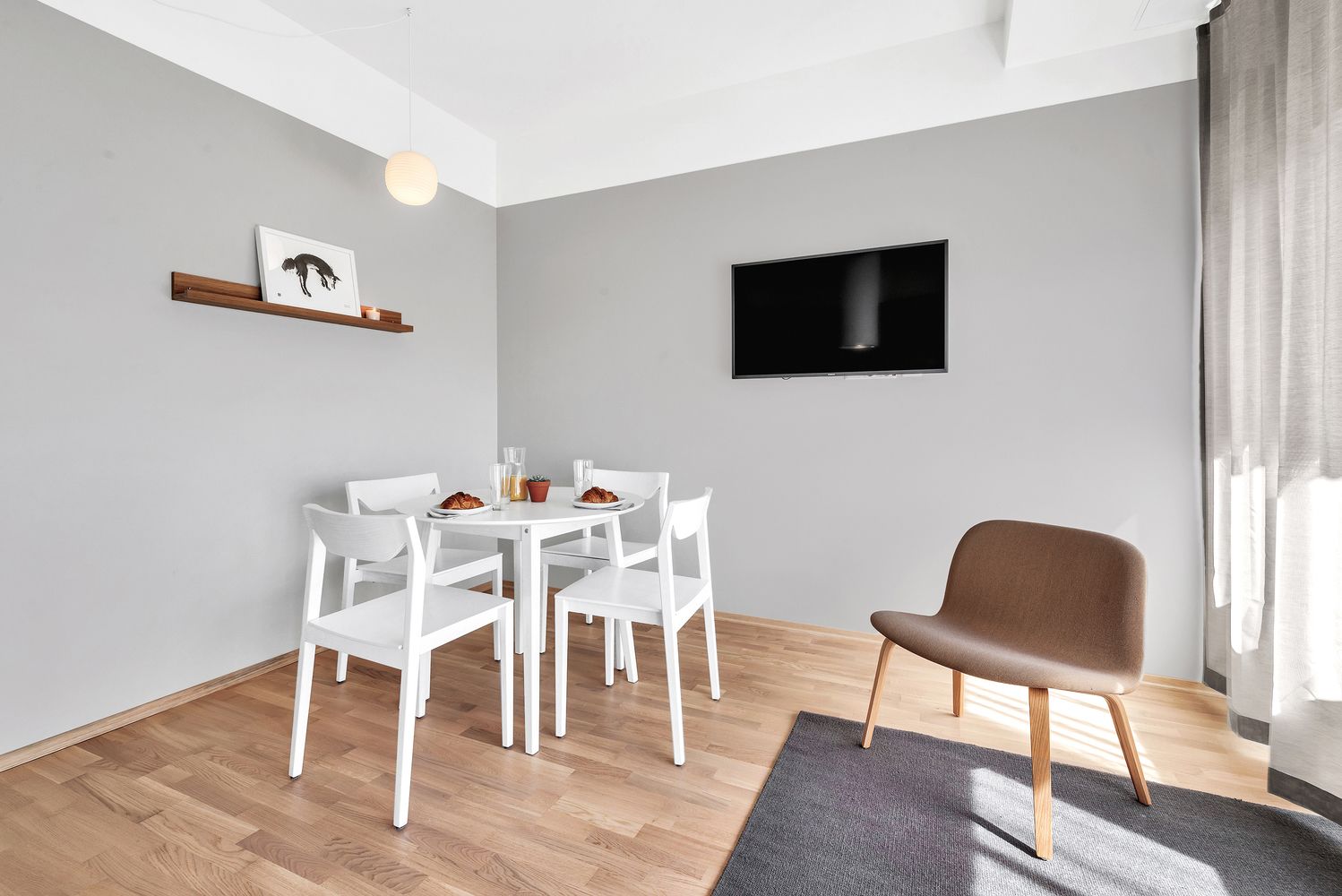 A modern living room with a dining table, white chairs, a brown armchair, a flat-screen TV, and a small shelf with a picture and two glasses.