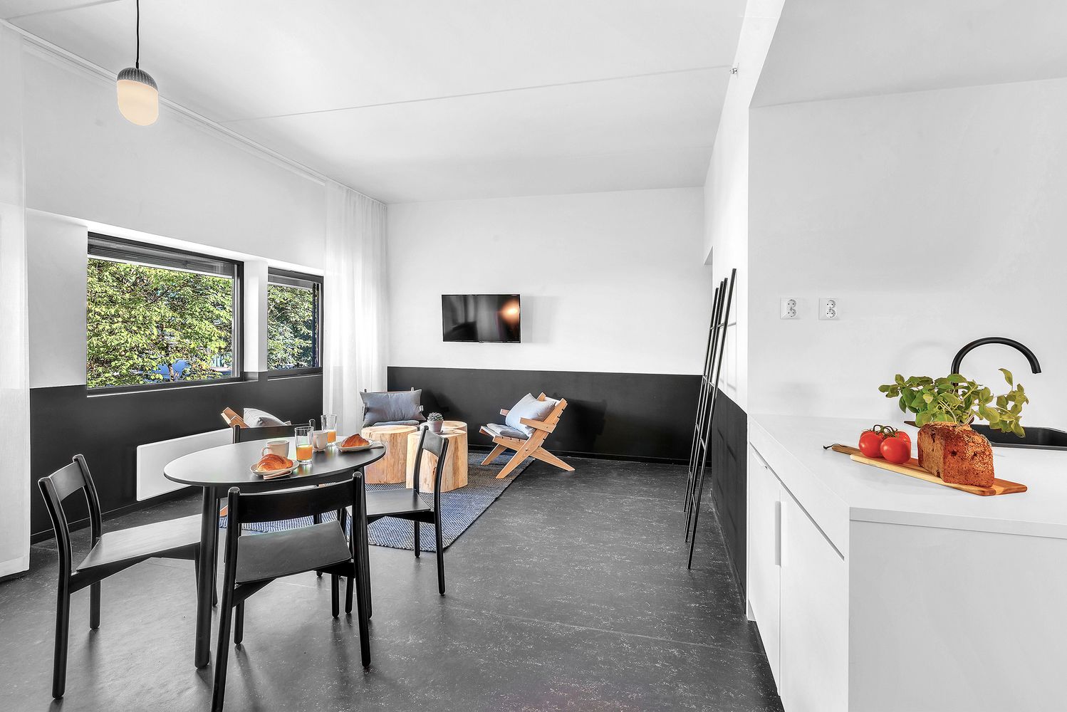 A modern kitchen with a dining area, a TV mounted on the wall, and a window with a view of greenery.