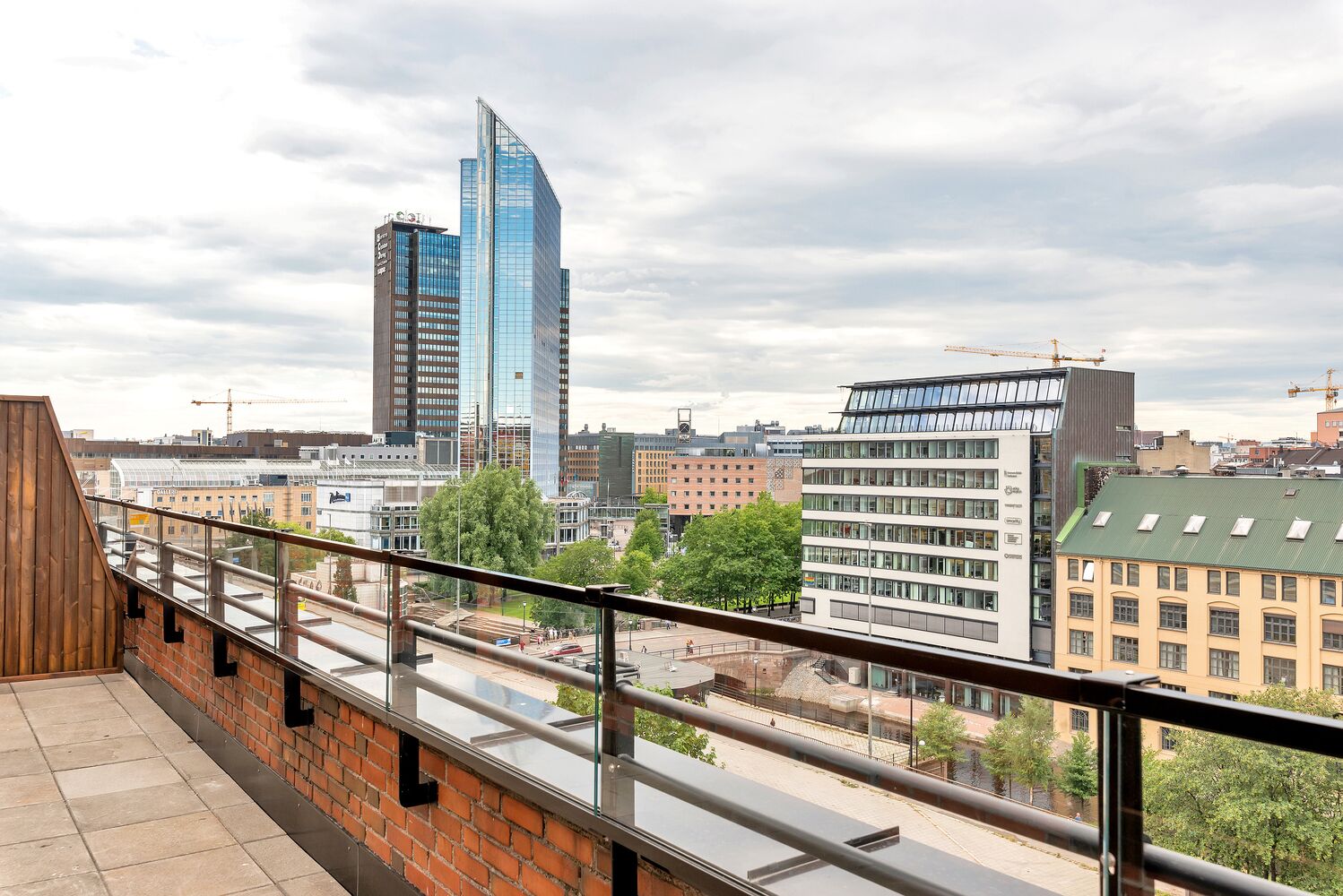 Un edificio alto con una facciata moderna in vetro, un muro in mattoni con ringhiera in metallo, e vista su un panorama urbano con diversi edifici e una gru edile sullo sfondo.
