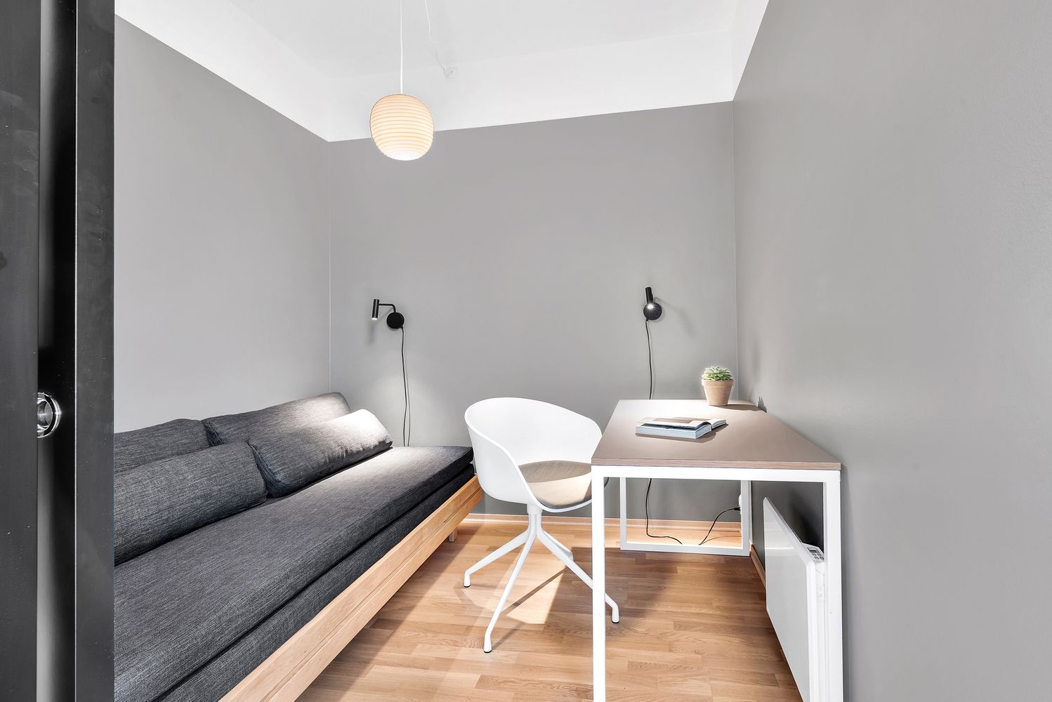 A modern, compact room with a grey sofa, a white chair, and a small desk with a plant.