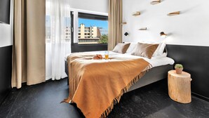 Habitación de hotel moderna con una cama grande, vista a edificios por la ventana y un taburete de madera con una planta.