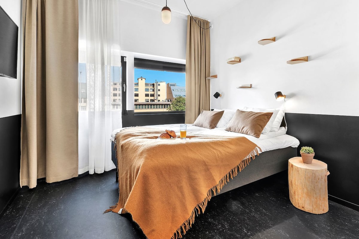 A modern hotel room with a large bed, a view of buildings through the window, and a small wooden stool with a plant.